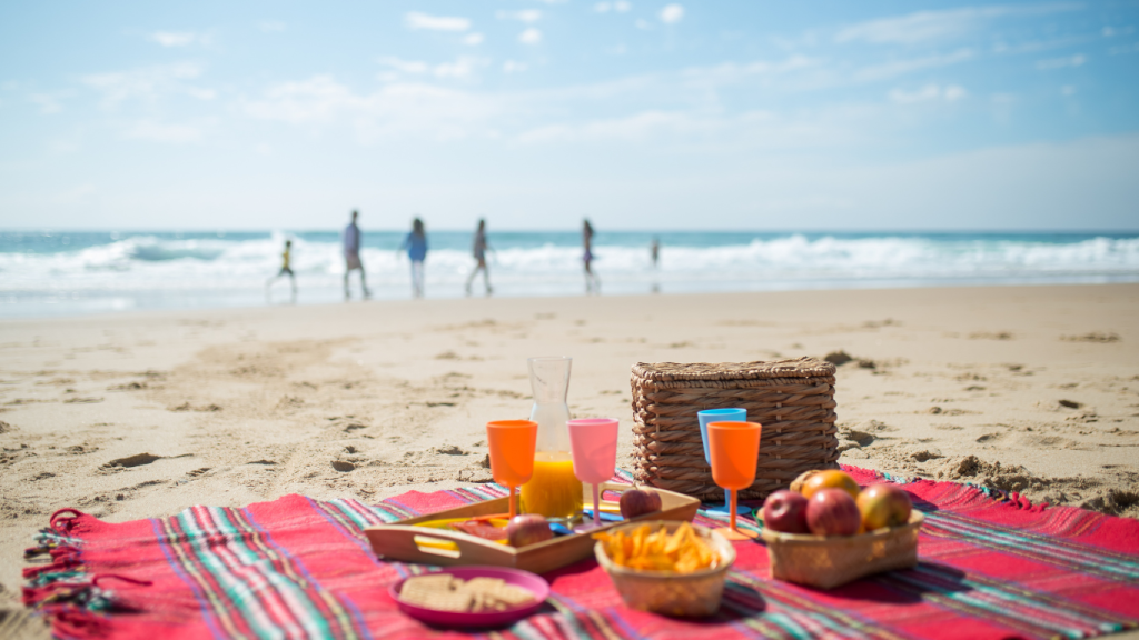 Na pierwszym planie piknik zero waste - czerwony koc i przekąski oraz napoje w kolorowych wielorazowych kubkach. W tle rodzina z dziećmi spacerująca po plaży.