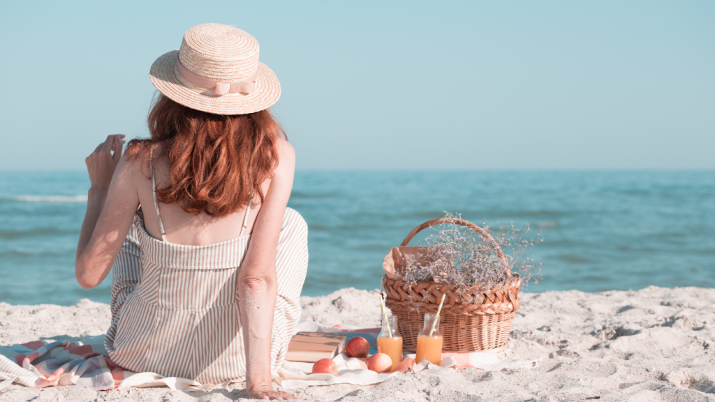 Kobieta w kapeluszu i letniej sukience siedzi na plaży i patrzy na morze. Obok niej piknik zero waste: koszyk i napoje w szklanych butelkach, książka.